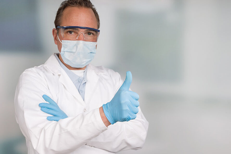 Mann mit Schutzkittel, Schutzbrille und OP-Maske zeigt Daumen nach oben.