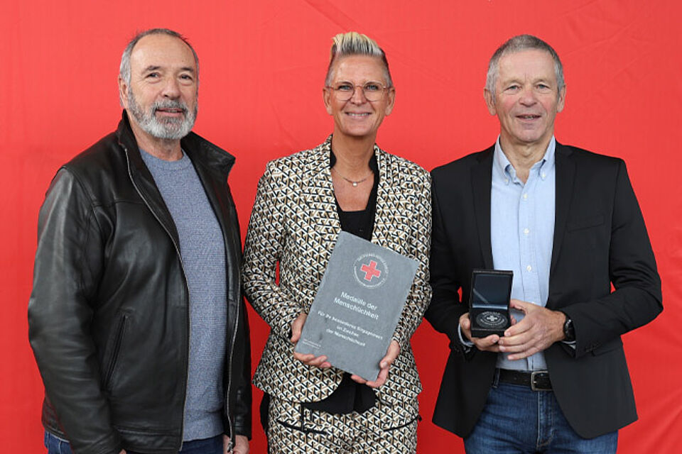 Zwei Männer und eine Frau halten die Medaille der Menschlichkeit