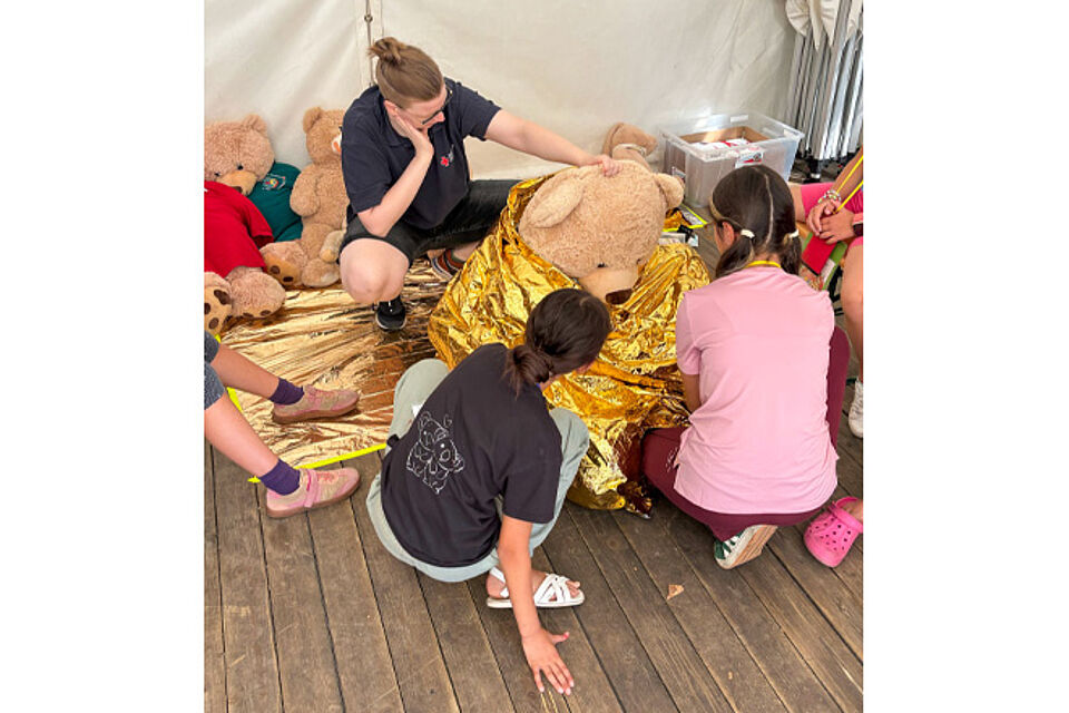 Ein Teddybär wird von zwei Kindern in eine Rettungsdecke gewickelt.