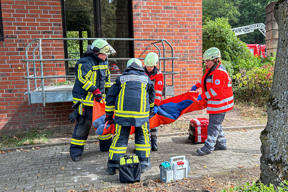 Zwei DRK-Helferinnen und zwei Feuerwehrleute tragen eine Puppe in einer Decke.