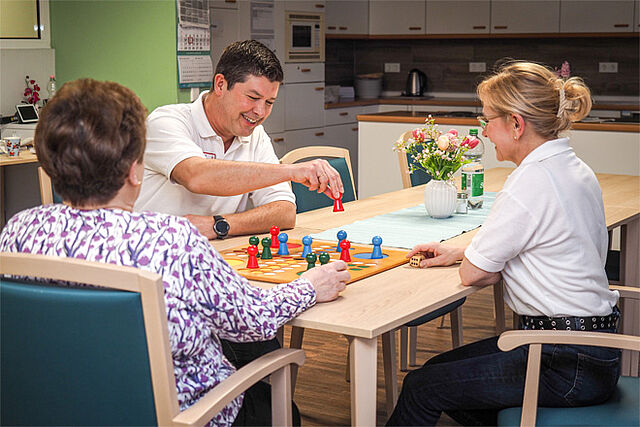 Zwei Pflegekräfte spielen mit einer Seniorin "Mensch ärgere Dich nicht".
