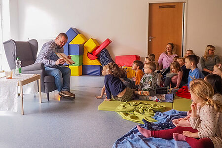 Ein Mann im Sessel zeigt Kindergartenkindern ein bebildertes Buch.