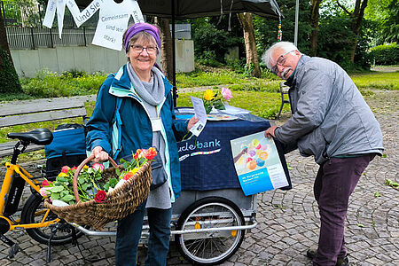 Ältere Frau mit Blumenkorb und älterer Mann stehen an einem Lastenrad.