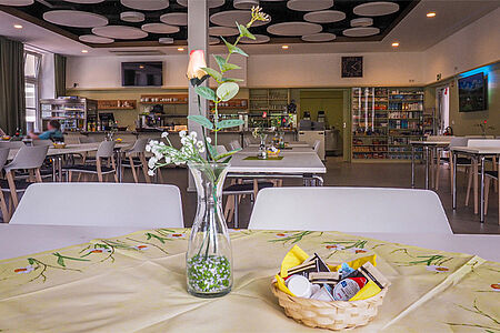 Blick in das Bistro des "Haus der Generationen. Im Vordergrund ein Tisch mit Blume im Hintergrund Tische und die Verkaufstheke.