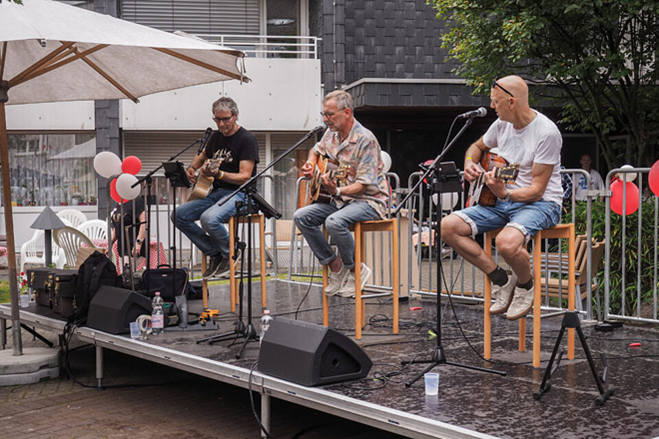 Drei Männer mit Gitarren sitzen auf Barhockern in einer Reihe und singen.