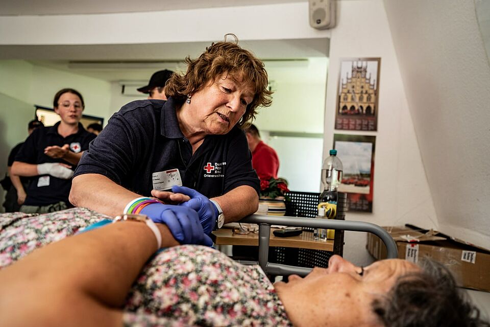 Frau mit DRK-Shirt kümmert sich um eine im Bett liegende Frau.