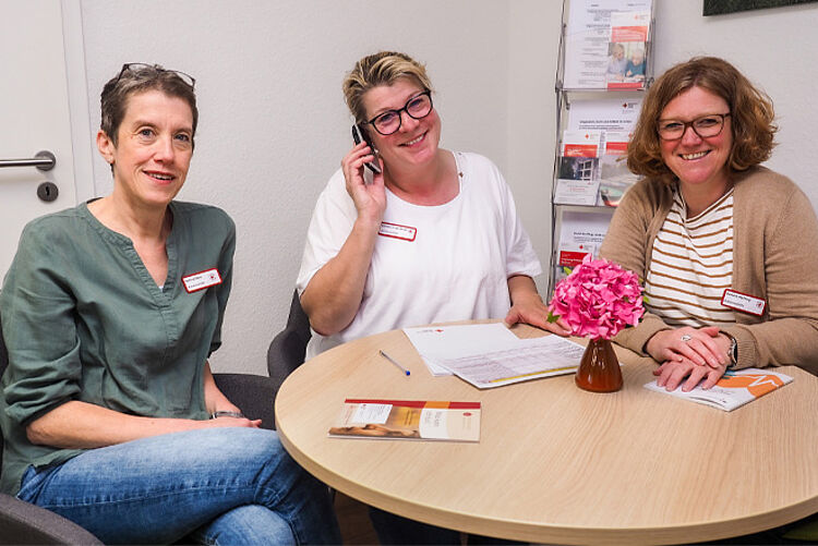 Drei Frauen sitzen an einem Tisch. Die Mittlere telefoniert.