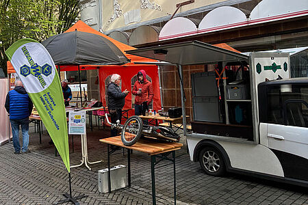 Ein gefaltetes Klappfahrrad liegt auf einer Bierbank vor einem kleinen Fahrzeug, dessen Seitenteile aufgeklappt sind und Werkzeuge freigeben.
