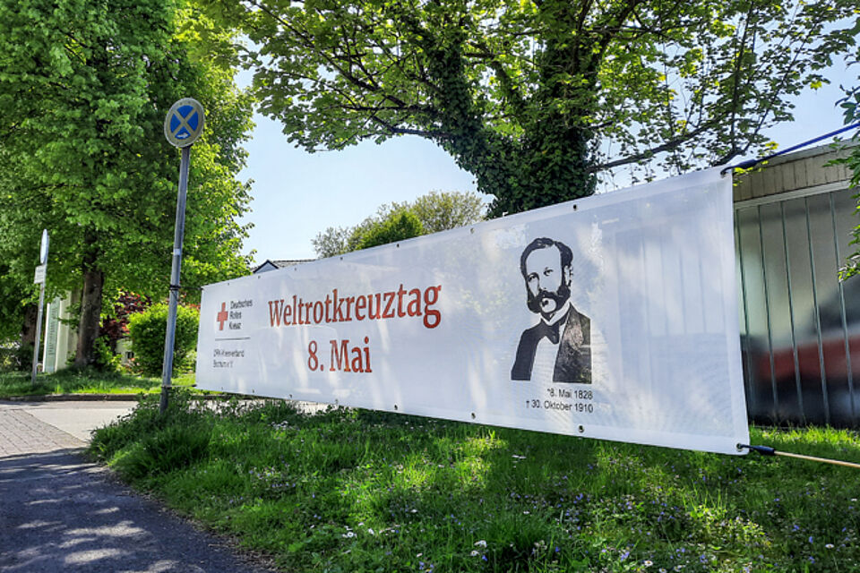 Ein Banner mit dem Schriftzug "Weltrotkreuztag 8. Mai" hängt vor einem grünen Baum.
