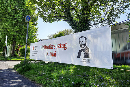 Ein Banner mit dem Schriftzug "Weltrotkreuztag 8. Mai" hängt vor einem grünen Baum.