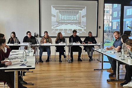Tagungsteilnehmer sitzen an Tischen in U-Form vor einer Beamer-Leinwand.