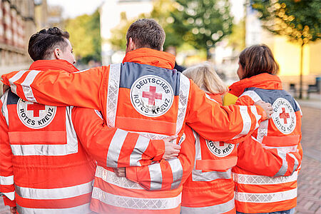 Vier Menschen in DRK-Jacken sind von hinten zu sehen, wie sie sich unterhaken.