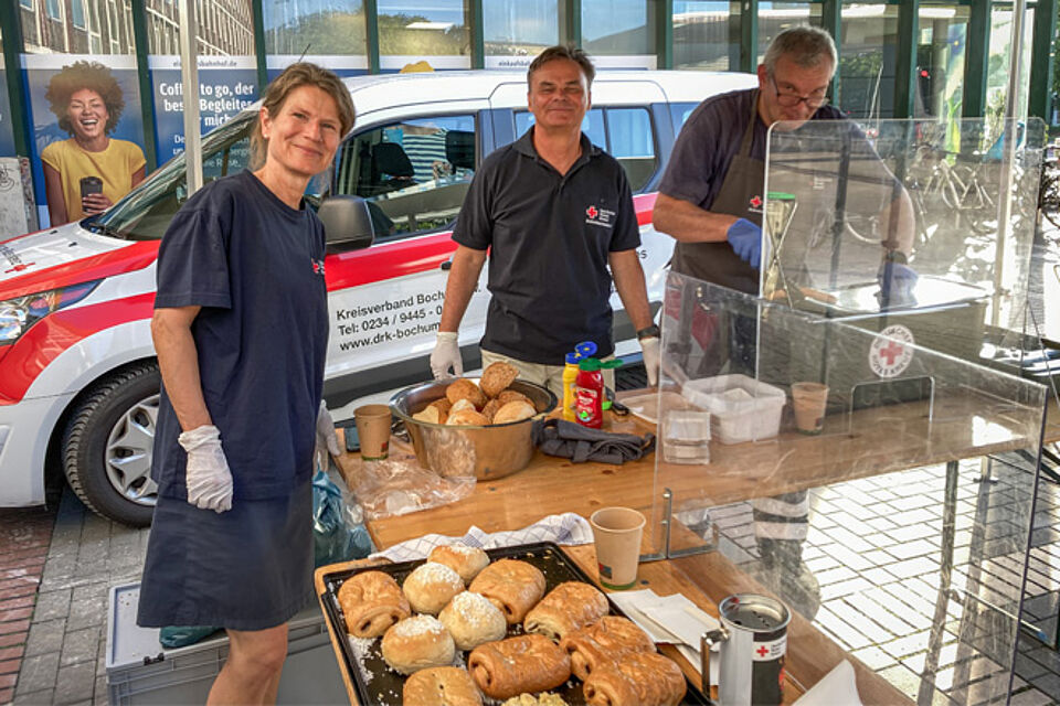 Eine Frau und zwei Männer in DRK-Kleidung stehen hinter einem Tisch mit Brötchen und Gebäck.