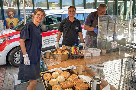 Eine Frau und zwei Männer in DRK-Kleidung stehen hinter einem Tisch mit Brötchen und Gebäck.