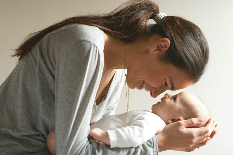 Eine junge Mutter hält ihr Baby vor sich auf den Unterarmen und berührt es mit der Stirn.