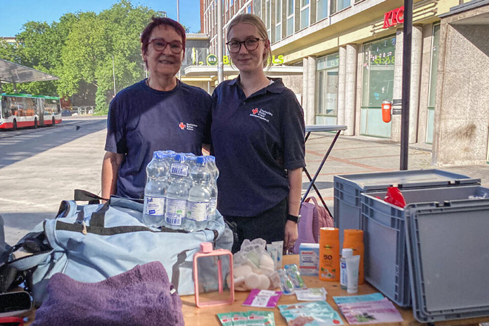 Zwei Frauen in DRK-Kleidung stehen hinter einem Tisch mit Schlafsack und Wasserflaschen.