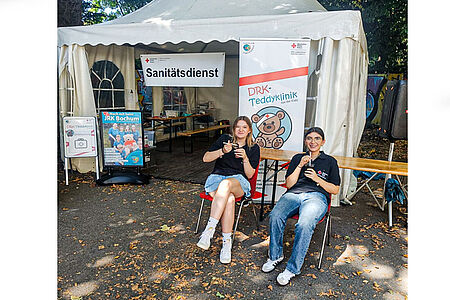 Zwei JRK-Helferinnen sitzen mit kühlen Getränken vor dem Pavillon der Teddyklinik.