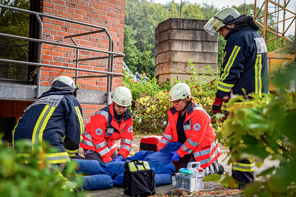 Zwei DRK-Sanitäter versorgen einen Verletzten, der von zwei Feuerwehrleuten vor einem Haus abgelegt wurde.
