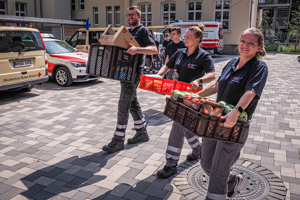 Ein DRK-Helfer und zwei -helferinnen tragen Verpflegungskisten.