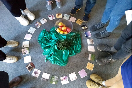 Teilnehmer stehen um kreisförmig ausgelegte Bierdeckel mit stimmungsvollen Motiven. In der Mitte ein grünes Seidentuch, darauf eine Schale mit Äpfeln und einem grünen Zweig.