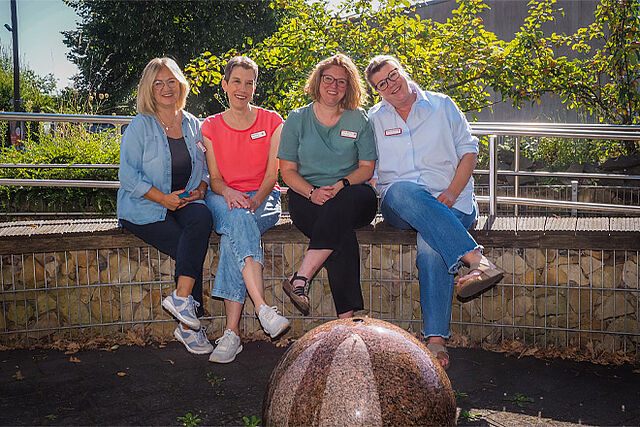Vier Damen mittleren Alters sitzen mit überschlagenen Beinen auf einer niedrigen Gartenmauer hinter einem kleinen Springbrunnen.