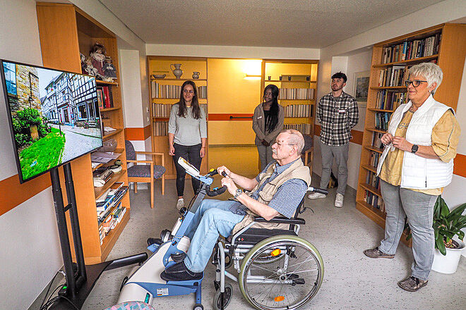 Ein Mann im Rollstuhl nutzt ein virtuelles Fahrradsystem: Strampeln vor einem Fernseher, der Routen zeigt.