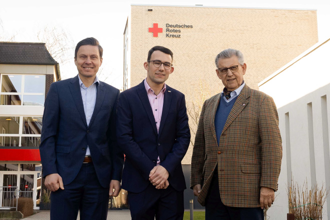 Drei Männer in Anzügen stehen vor einem Gebäude mit "DRK"-Logo.