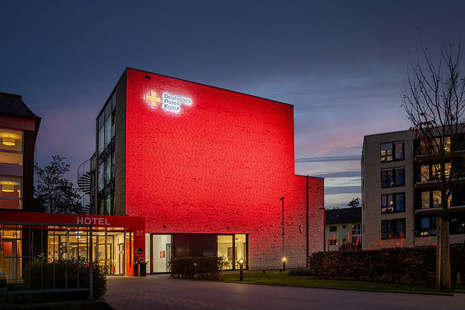 Ein rot beleuchtetes, dreistöckiges Gebäude mit fensterloser Front und DRK-Logo.
