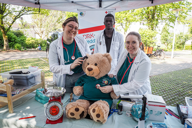 Zwei Frauen und ein Mann in weißen Kitteln untersuchen einen großen Teddybären.